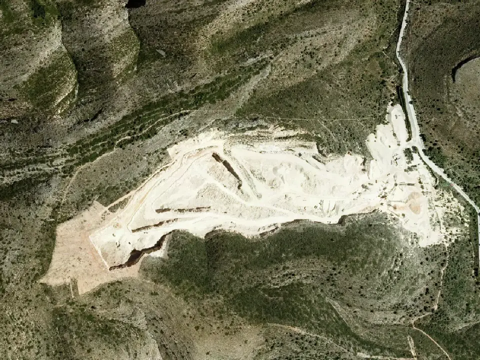 Ortofoto de una explotación a cielo abierto en ladera, con la zona excavada en blanco rodeada de monte bajo y una carretera a la derecha