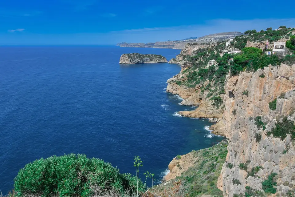Acantilados de la costa de Xàbia con pinos y casas en lo alto y un islote frente a la orilla